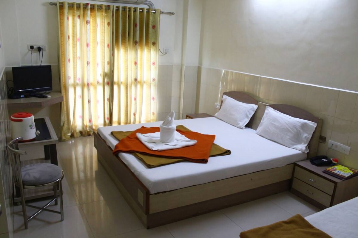 Quarto de hotel com cama, uma escrivaninha com computador, uma cadeira e janela com cortinas.