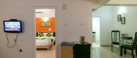 Habitación de hotel con cama, escritorio, silla, televisor y zona de comedor con sillas y mesa.