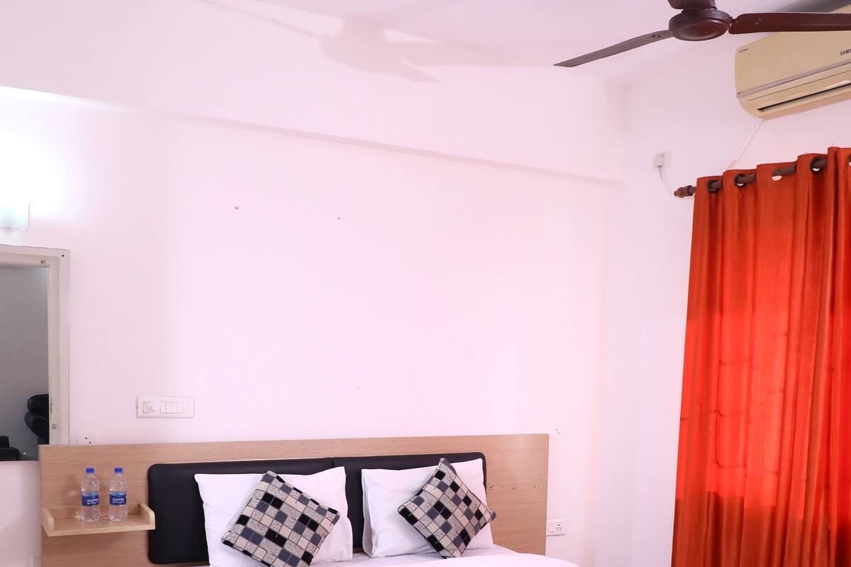 A neatly made bed with a checkered blanket, a wooden headboard, and two pillows. There is a nightstand with a water bottle and a mirror. A ceiling fan is visible above the bed.