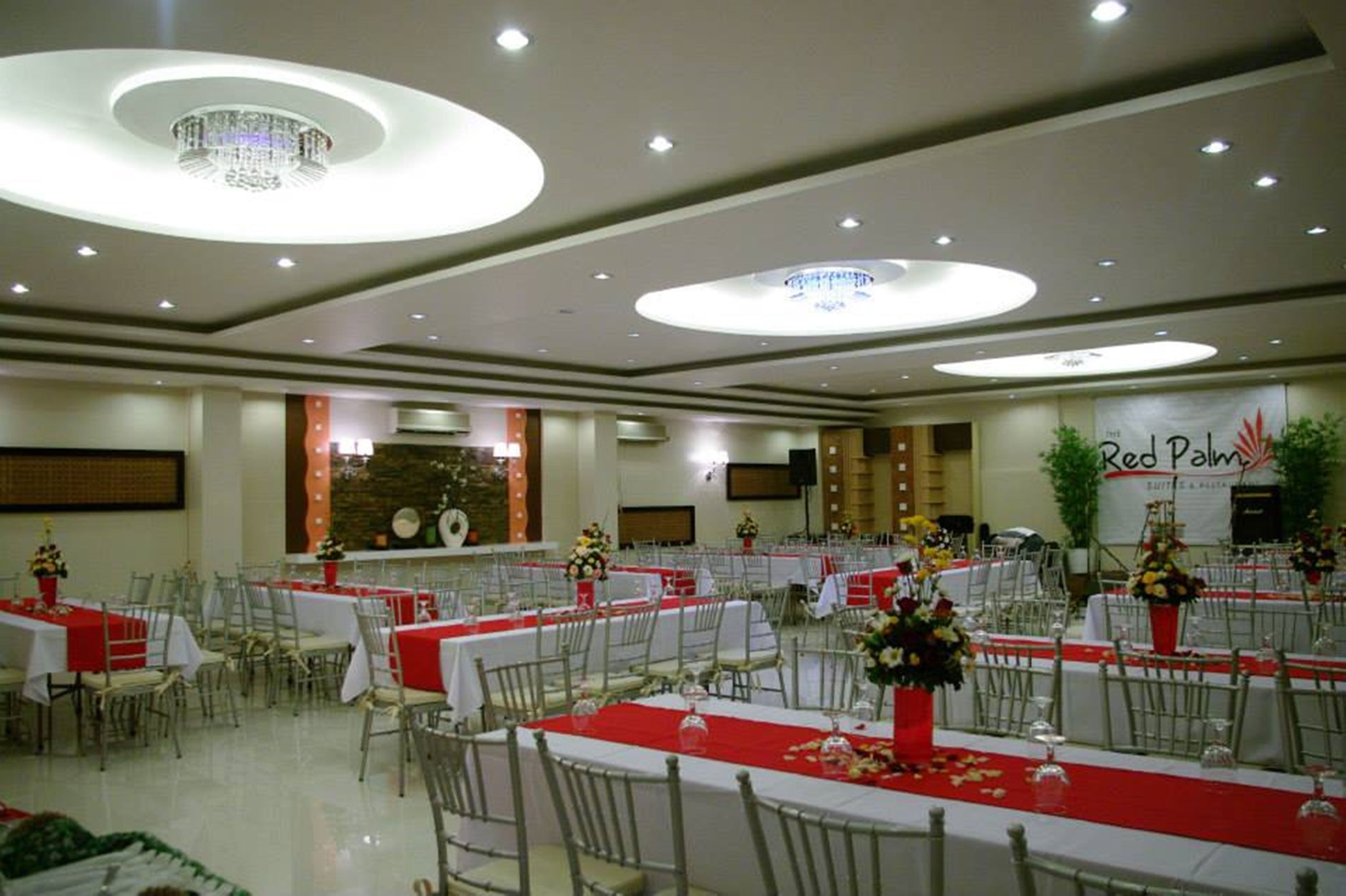Un salón de banquetes con mesas redondas, manteles rojos y sillas blancas.