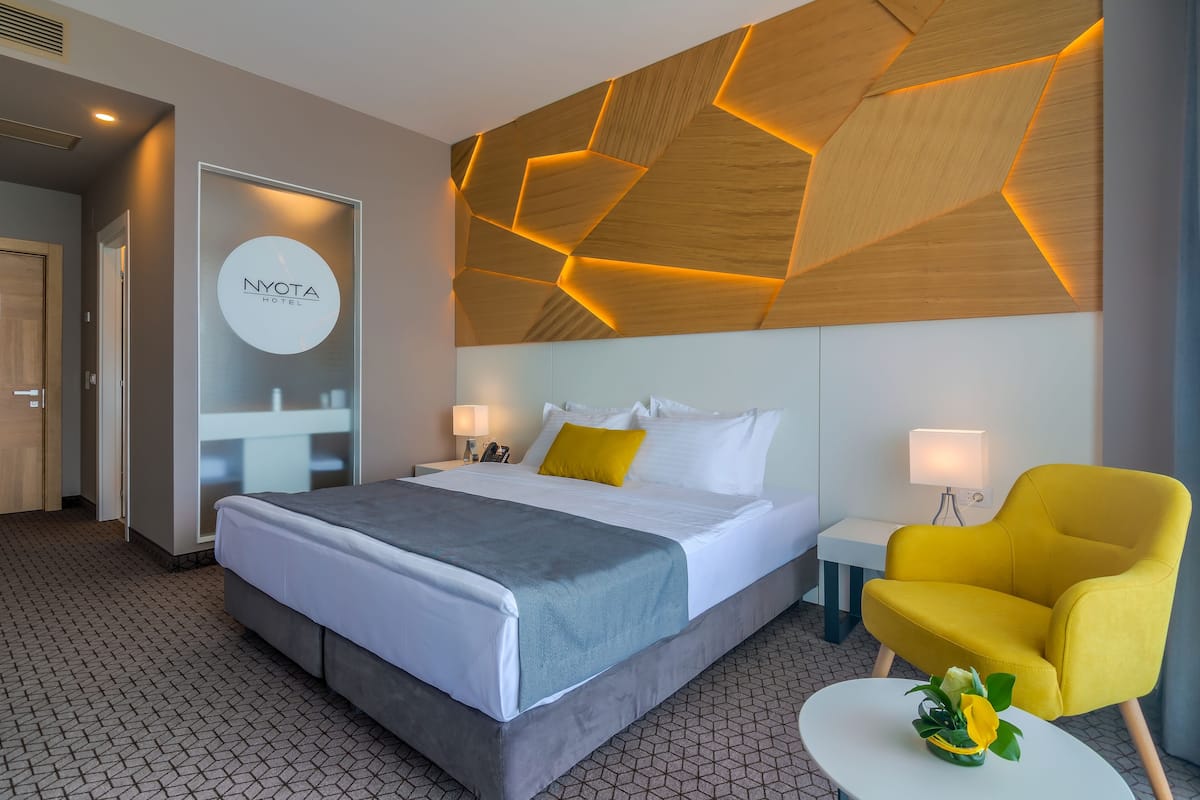 A hotel room with a bed, a yellow armchair, a small table with a plant, and a geometric wooden ceiling design.