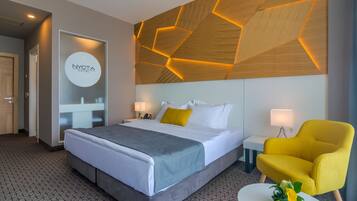 A hotel room with a bed, a yellow armchair, a small table with a plant, and a geometric wooden ceiling design.