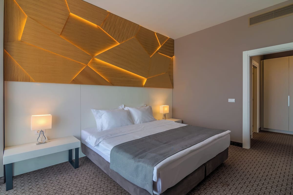 A modern hotel room with a large bed, bedside tables with lamps, a geometric patterned ceiling, and a wardrobe.