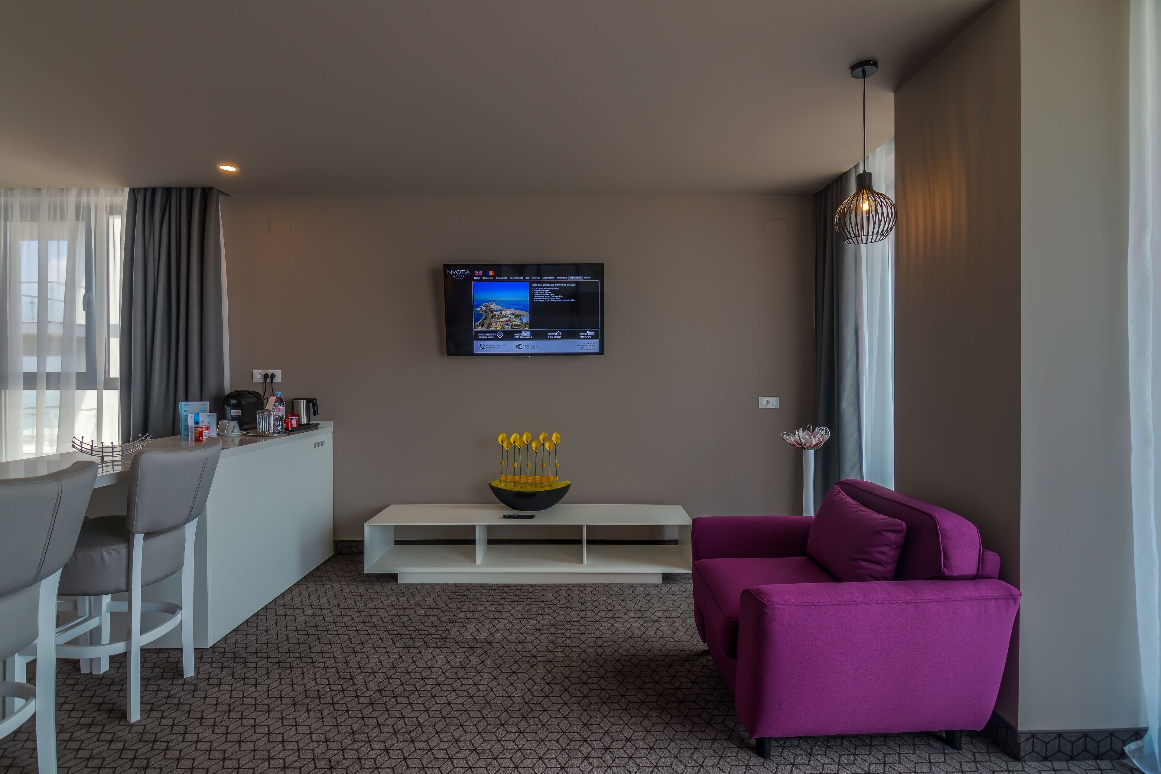 A modern living area with a flat-screen TV mounted on the wall, a white console table, a purple sofa, and a dining area with bar stools.