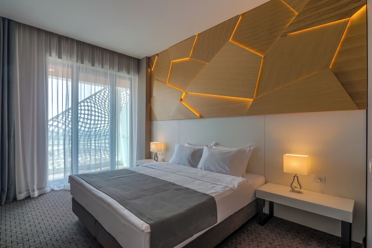 A modern hotel room with a large bed, two bedside tables with lamps, a geometric-patterned wall, and a view through large windows.