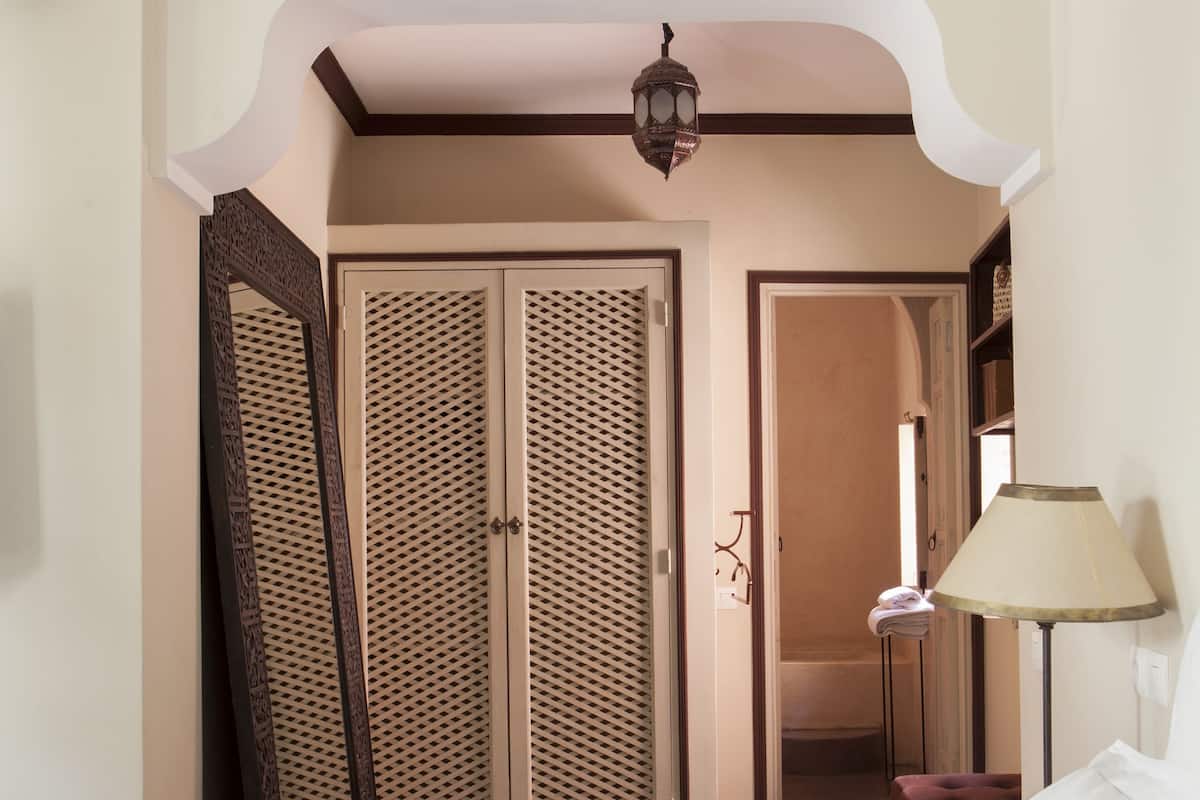 A hotel room with a bed, a bedside table with a lamp, a tray with glasses and a bowl, a woven hat, and a wardrobe with a mirror.