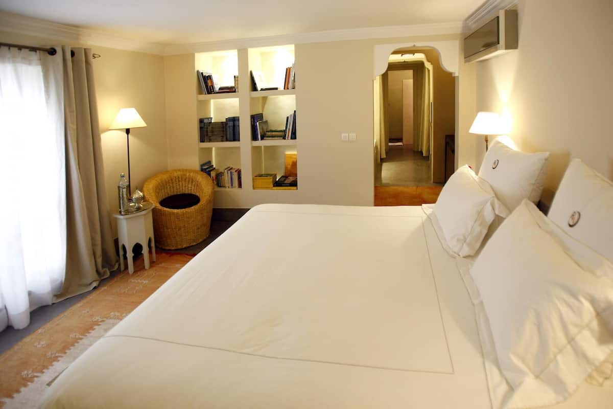 A hotel room with a large bed, two bedside lamps, a wicker chair, a bookshelf, and a mirror.