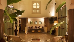 Un área de descanso acogedora con un conjunto de sofás, una mesita y sillas. Hay plantas grandes y un jarrón decorativo sobre la mesa.