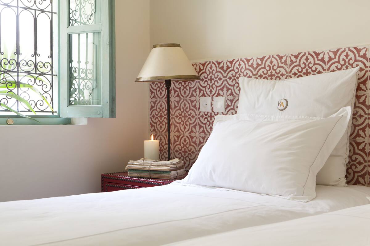 A neatly made bed with white linens, a bedside lamp, a stack of books, and a lit candle on a nightstand.