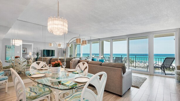 Un vaste espace de vie agrémenté d’une table en verre, de chaises blanches et d’un grand canapé. Un lustre est suspendu au plafond et la pièce est dotée de grandes fenêtres offrant une vue sur la plage.