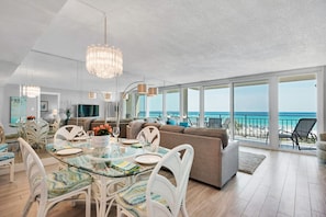 A spacious living area with a glass table, white chairs, and a large sofa. A chandelier hangs from the ceiling, and the room has large windows offering a view of the beach. - The Breakers of Fort Walton Beach (Fort Walton Beach)
