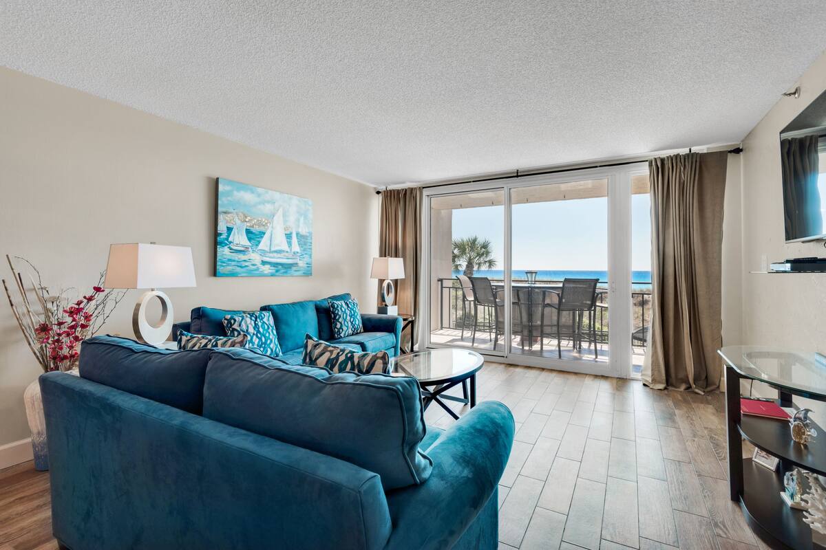 A modern living room with a blue sofa, a glass coffee table, and a painting of sailboats. There is a balcony with a view of the sea.