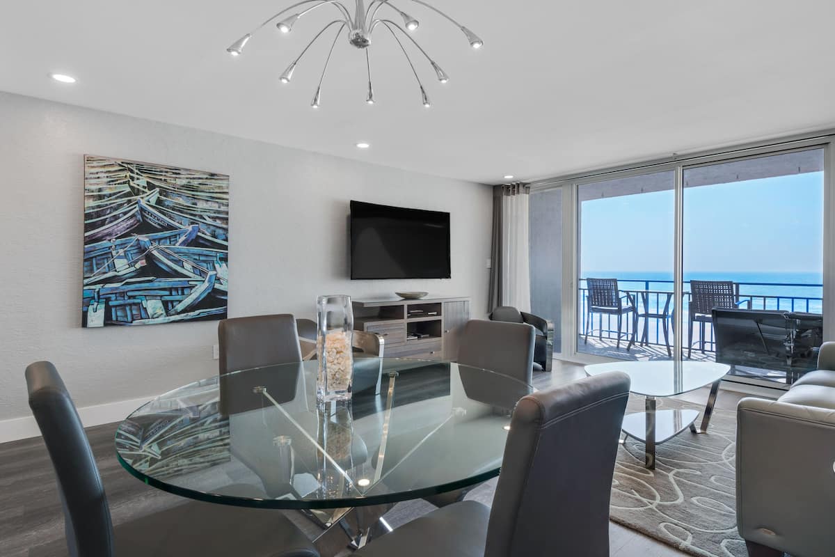 A modern dining area with a glass table, leather chairs, a flat-screen TV, and a painting of boats on the wall.