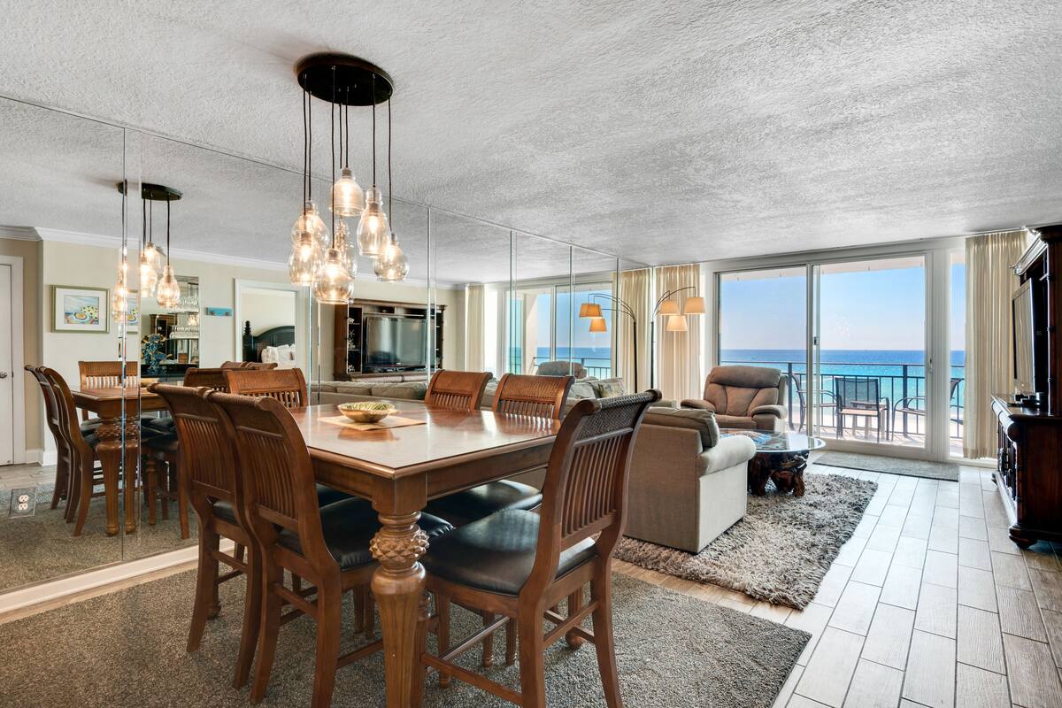A spacious living area with a large dining table, multiple chairs, and a sofa. There is a television and a kitchen area in the background.