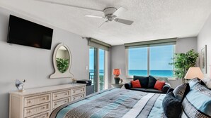 Une chambre à coucher comprenant un lit, une commode, un téléviseur, un ventilateur de plafond, une fenêtre donnant sur l’océan et une plante.