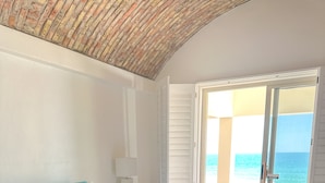 Une chambre avec un grand lit, une table de chevet, une lampe et une vue sur l’océan par une porte ouverte.