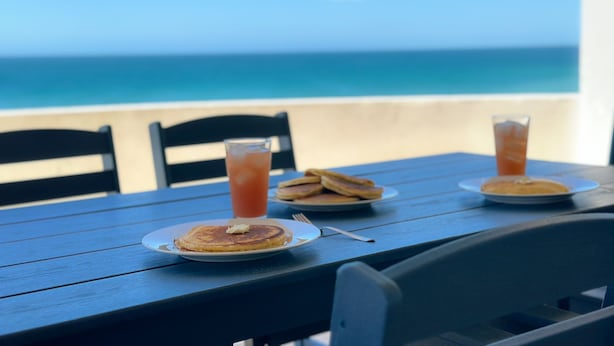 Une table dressée avec des aliments pour le petit-déjeuner, notamment des crêpes et des boissons glacées, donnant sur une plage.