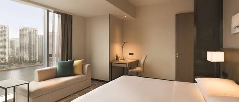 A hotel room with a large bed, a sofa, a desk, and a chair. There is a window with a view of buildings and a lamp on the desk.