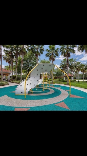 Explore the fun of this playground with a climbing wall and slide, surrounded by a vibrant patterned ground and lush palm trees.