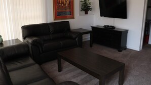 A living room with a black leather sofa, a wooden coffee table, a small black side table, a potted plant, and a mounted flat-screen TV.