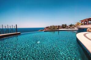 Uma piscina azul cristalina com vista para o oceano, um deck de madeira e um edifício com arcos.