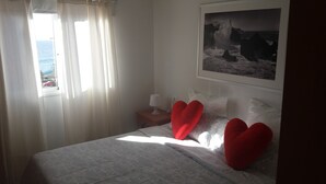 Quarto com cama, mesa de cabeceira, abajur, almofadas em formato de coração, janela com cortinas e um quadro na parede.