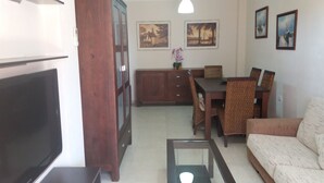 Uma sala de estar com televisão, mobiliário de madeira, um sofá e um espaço de jantar com cadeiras e uma mesa.