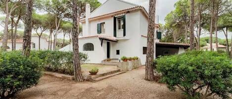 A white villa with a tiled roof, surrounded by trees and a gravel driveway.