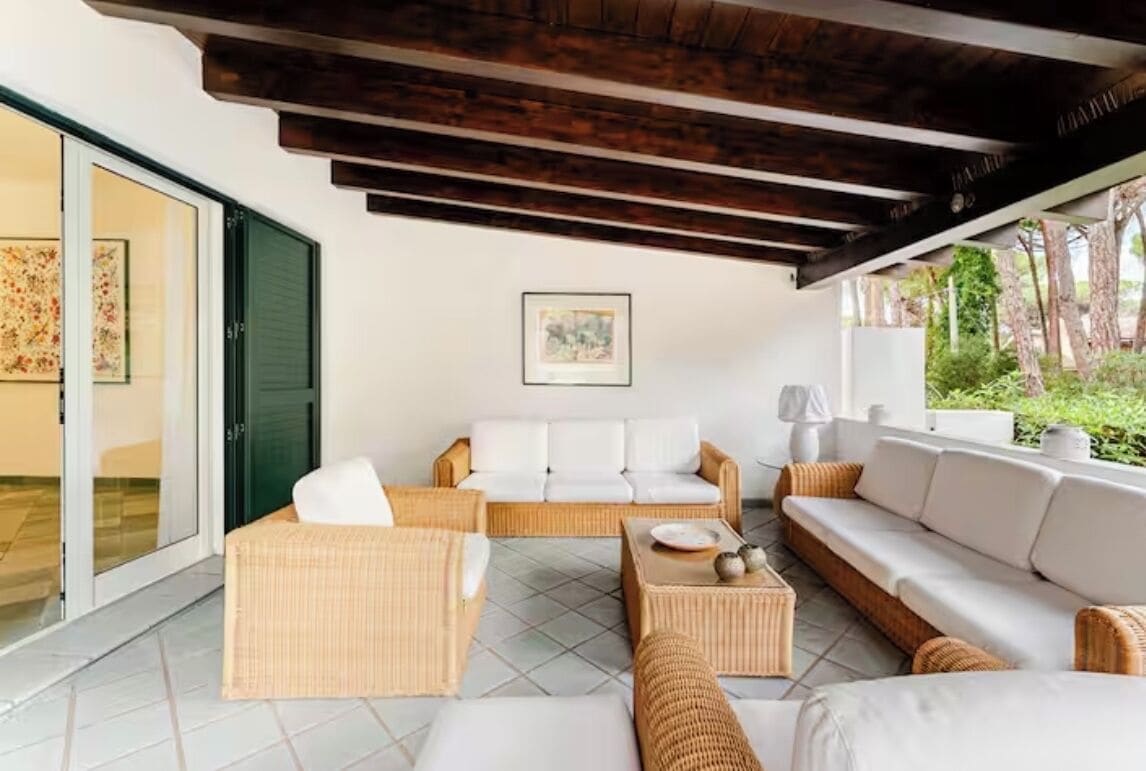 Una sala de estar moderna con mobiliario de mimbre, techo de madera y un amplio ventanal con vistas a la vegetación.