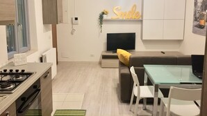 A modern kitchen with a gas stove, a dining area with a glass table and chairs, a sofa with a yellow pillow, a TV, and a wall-mounted sign that says "Smile".