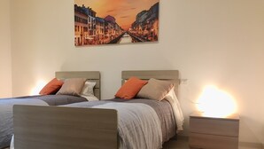 A bedroom with two beds, a nightstand with a lamp, a radiator, and a painting of a cityscape on the wall.