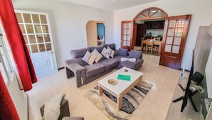 A living room with a grey sofa, a coffee table, and a TV. There is a dining area with a wooden table and chairs. - Casa da Achada in Arco da Calheta (Arco da Calheta)