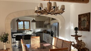 A dining area with a wooden table and woven chairs, a chandelier, and a view into the kitchen. - Provencal country house with heated freeform pool (Châteaurenard)