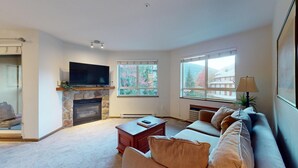 Ein Wohnzimmer mit Kamin aus Stein, einem Flachbildfernseher, einer Couch und zwei Fenstern mit Blick auf Bäume und Gebäude.