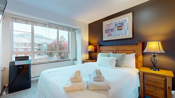 Ein Hotelzimmer mit einem großen Bett, zwei Nachttischlampen, einem Holznachttisch, einem gerahmten Bild an der Wand und einem Fenster mit Blick auf Gebäude und Bäume.