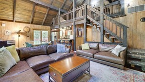 A rustic cabin interior with a loft bedroom, a dining area, and a living room with leather sofas and a wooden coffee table.