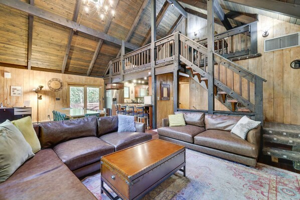 A rustic cabin interior with a loft bedroom, a dining area, and a living room with leather sofas and a wooden coffee table.