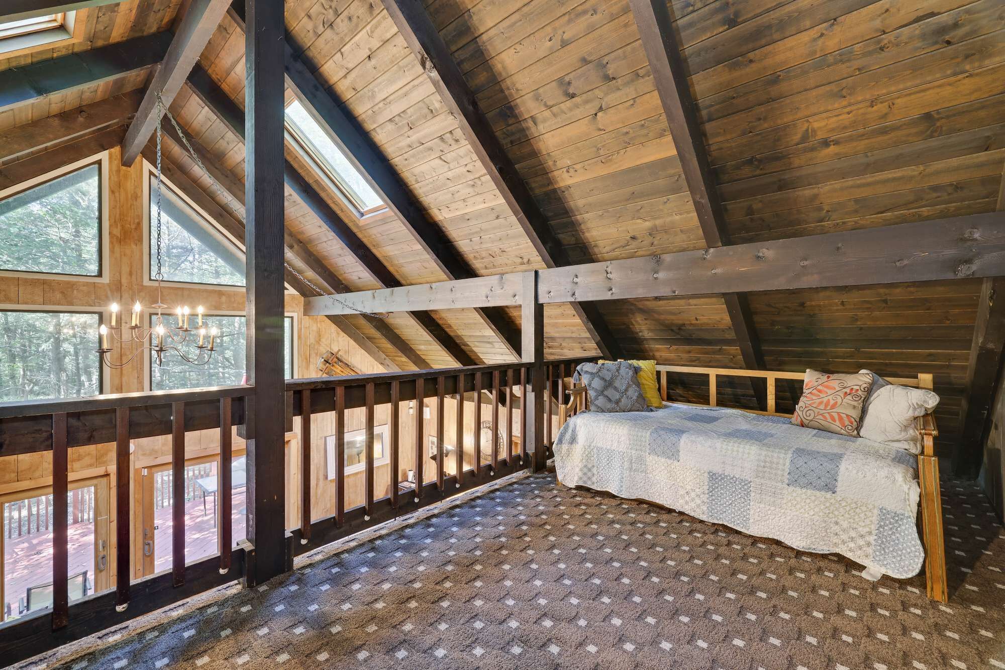Un dormitorio acogedor en el ático, con techo de madera, cama con ropa de cama estampada y ventanas amplias que ofrecen vistas al exterior.