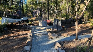 A gravel path bordered by rocks, leading through a wooded area with trees and a rustic cabin in the background.