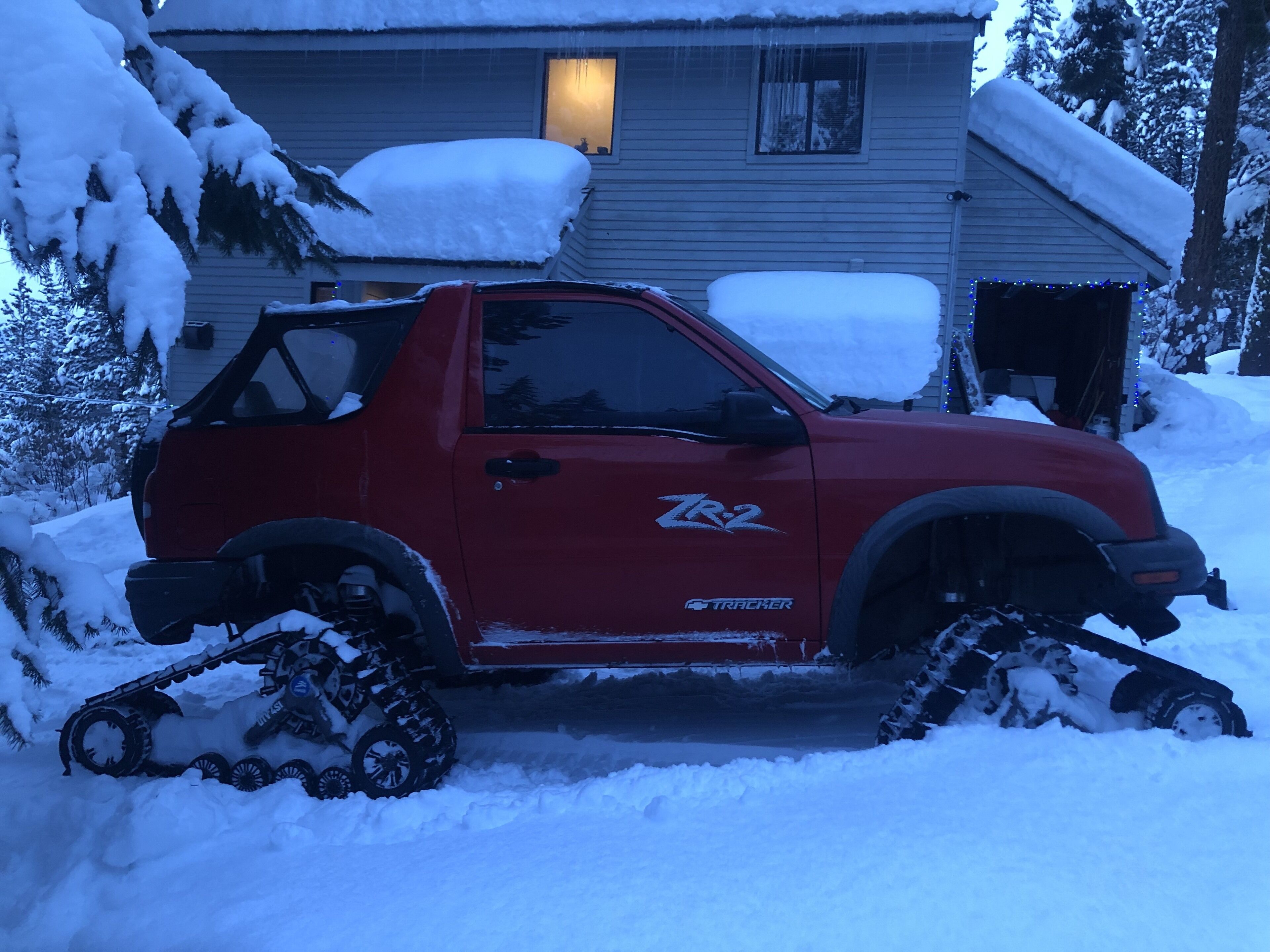 A red pickup truck with snow tracks, parked in front of a house with snow on the roof and windows.