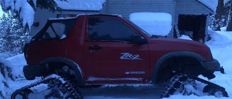A red pickup truck with snow tracks, parked in front of a house with snow on the roof and windows.