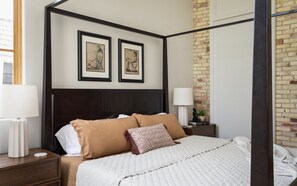 Uma cama com dossel e um painel de cabeceira escuro, duas obras de arte emolduradas acima e uma parede de tijolos à direita.