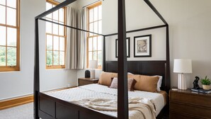 A four-poster bed with a dark wooden frame, a white bedspread, and beige pillows. There are two bedside tables with lamps and a small plant. The room has large windows with curtains, and a framed picture on the wall. - Historic Hearth · Newly Redesigned Condo in Historic Building (Traverse City)