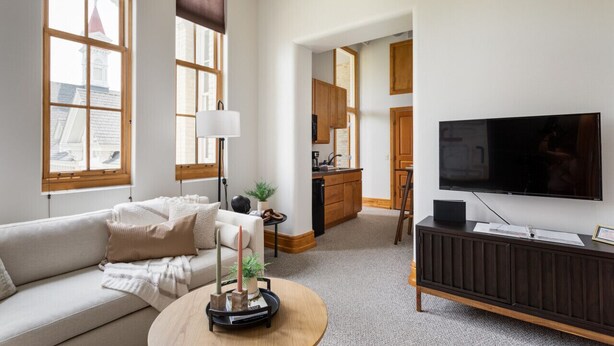 A modern living room with a sofa, a coffee table, and a TV on a wooden stand. There is a kitchen area with wooden cabinets and a window with a view of a building. - Historic Hearth · Newly Redesigned Condo in Historic Building (Traverse City)