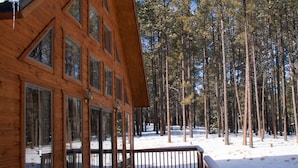 Una cabaña de madera con ventanas grandes, una terraza y un bosque nevado al fondo.
