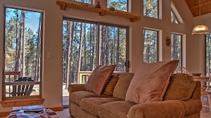 Una sala acogedora con un sofá marrón, ventanas amplias y vistas a un bosque.