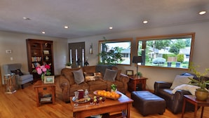 A spacious living room with a brown leather sofa, a wooden coffee table with fruit and drinks, a bookshelf, and large windows with a view of the backyard. - HEATED Pool & Hot Tub in a Quiet Neighborhood- Walk to Town- Dog Friendly (Walla Walla)