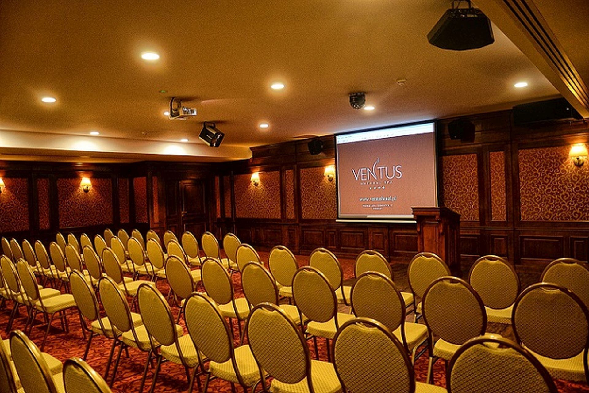 A conference room with rows of chairs, a podium, and a projection screen displaying "VENTUS".