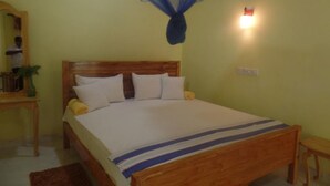 A single bed with a wooden headboard, white bedding, and a blue-striped blanket. There is a small bedside table with a plant and a wall-mounted light fixture. - Peace Heaven (Udawalawa)