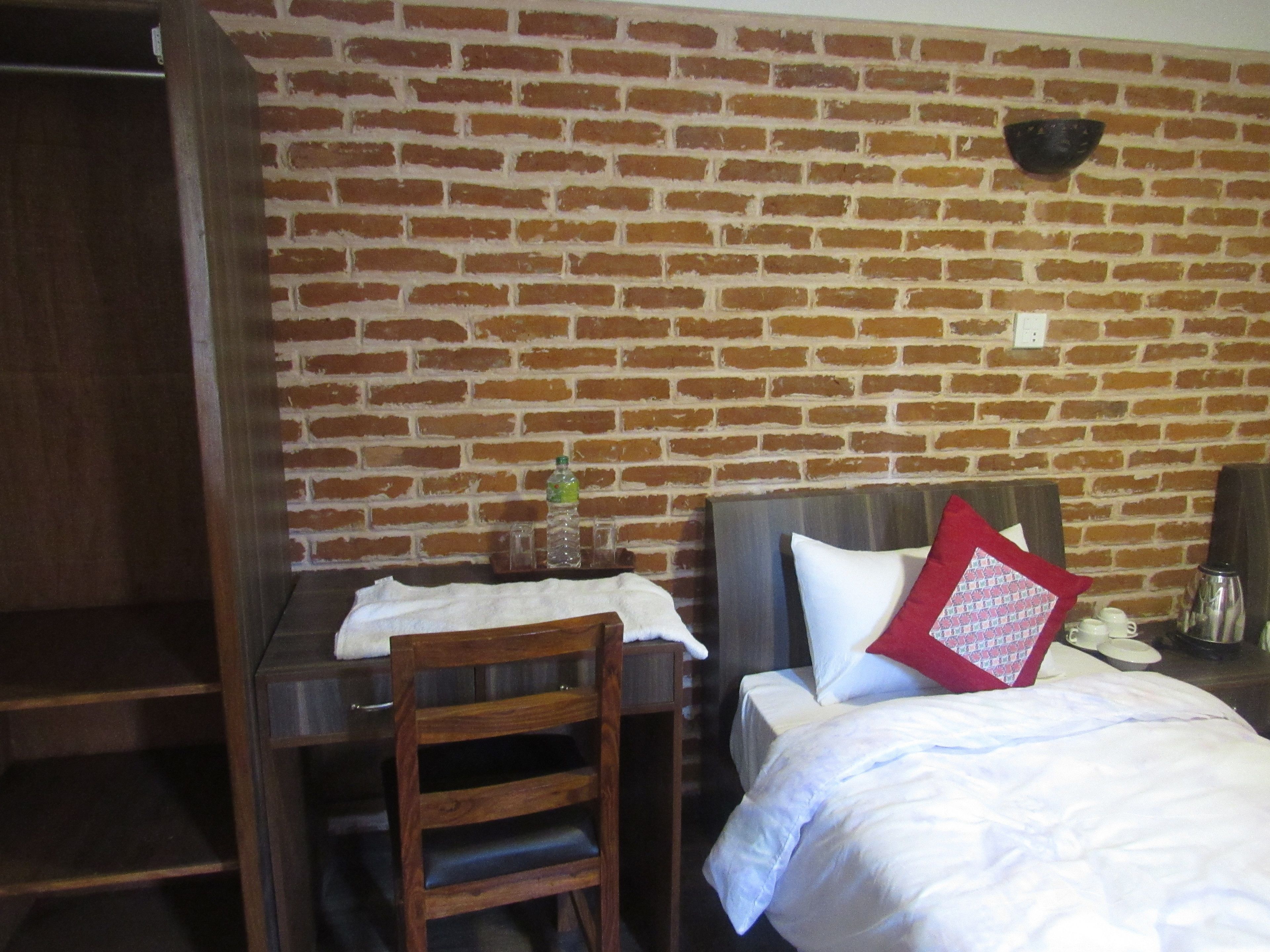 Una habitación con pared de ladrillo, un escritorio de madera, una silla, una cama con ropa blanca y una almohada roja, y un pequeño estante con una botella y vasos.